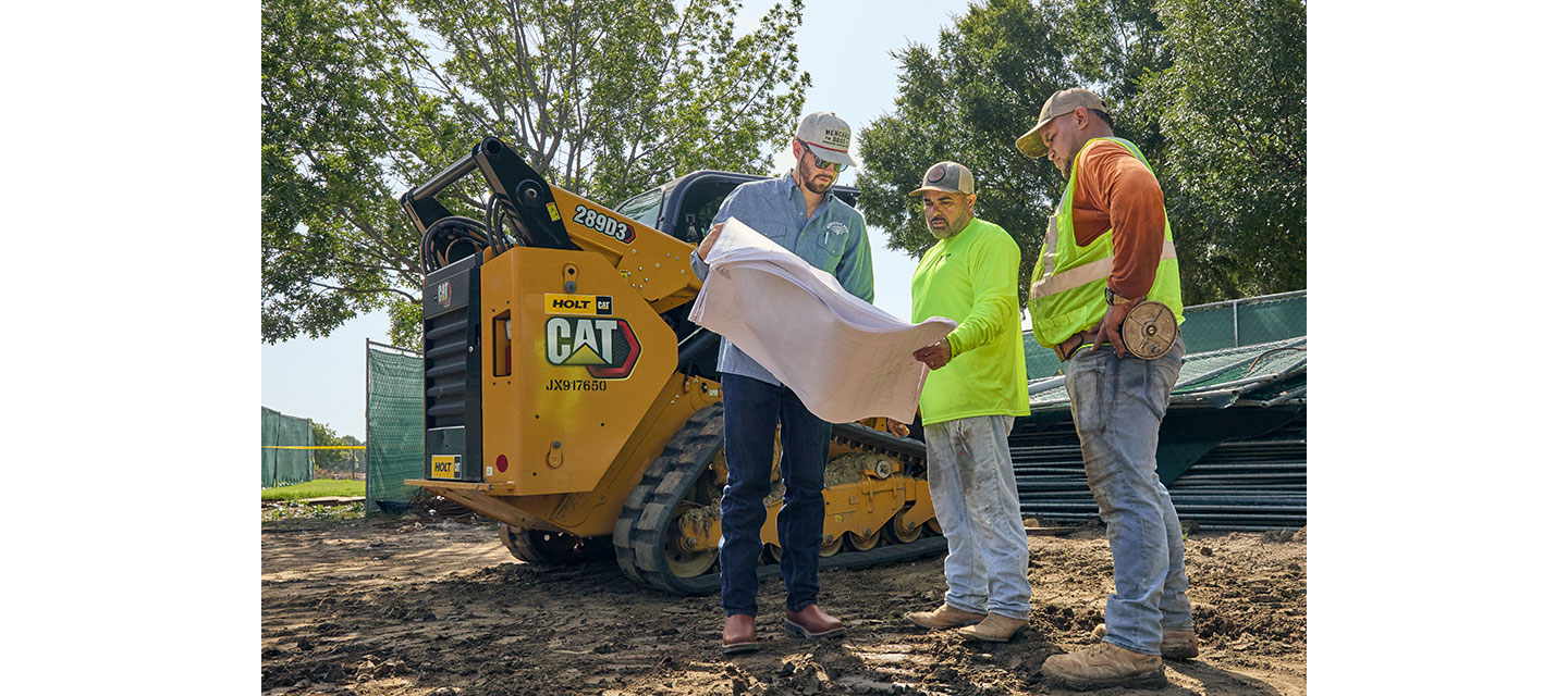 Three men, Nathan Menchaca and two workers, review blueprints at a job site beside a yellow CAT skid steer loader, with safety gear and work boots visible on a dirt-covered ground.